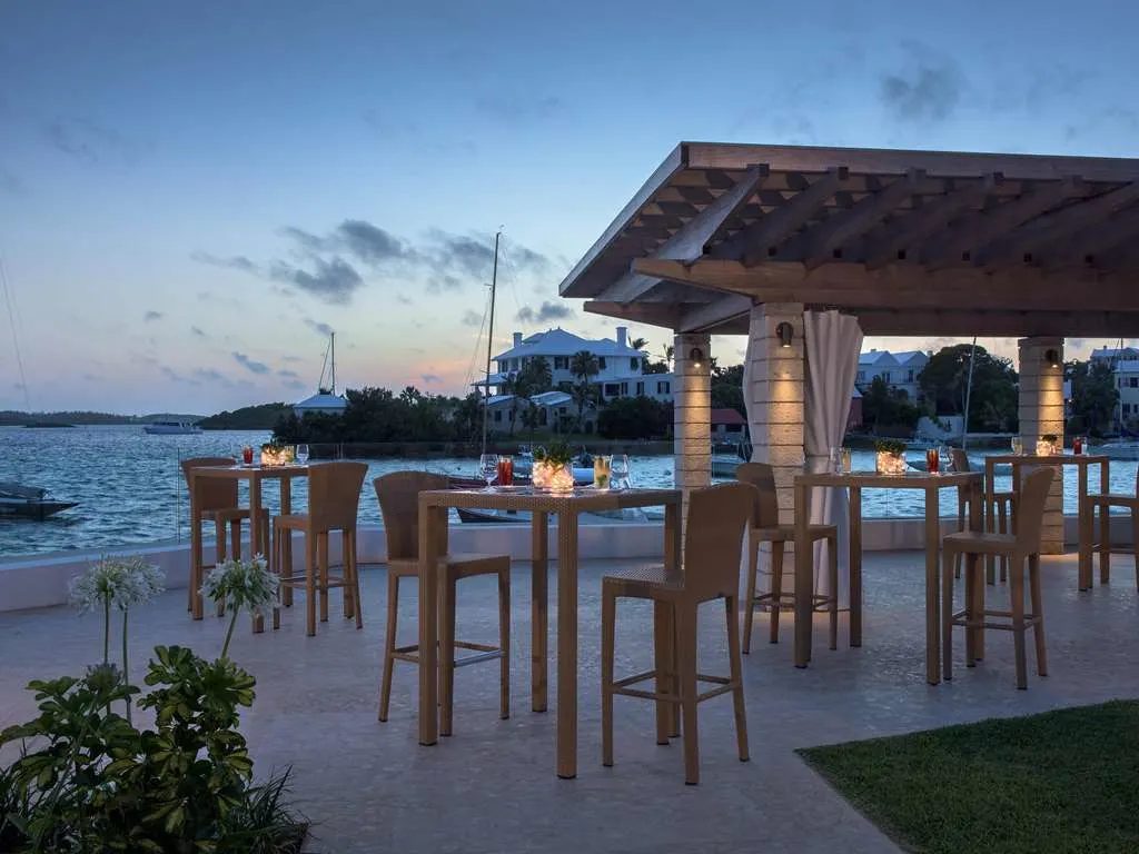 Candlelit high-top tables and a pergola bar by the water at dusk