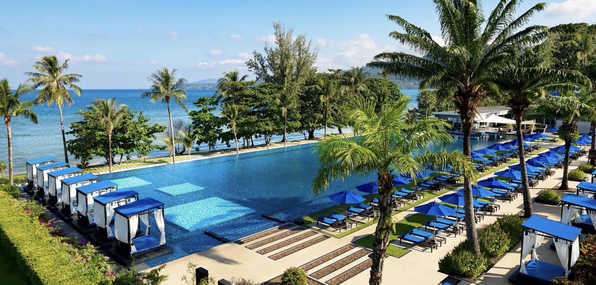 A long blue pool lined with cabanas and palms beside the Andaman Sea