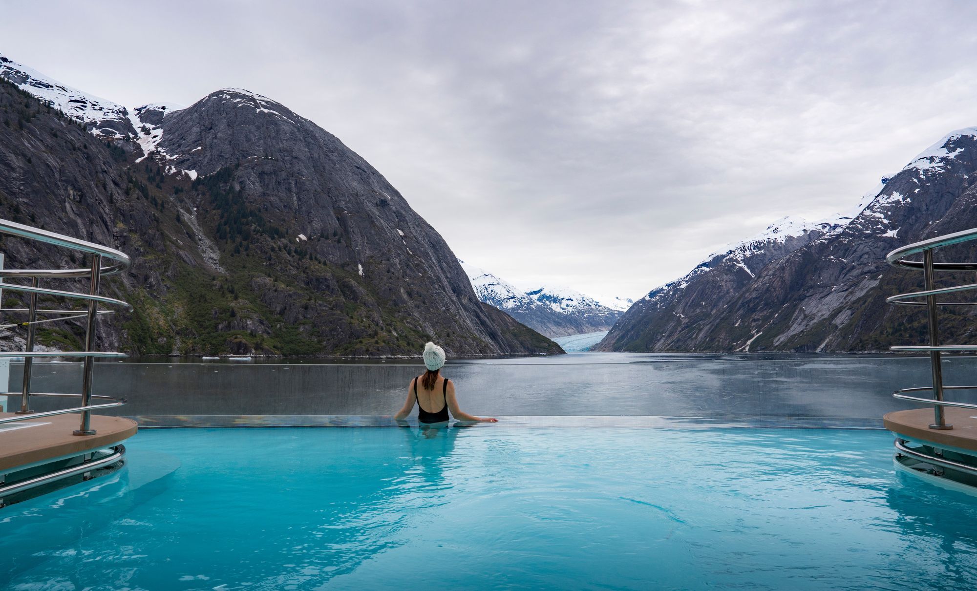 A guest in an infinity pool aboard ship, facing the mountains near Dawes Glacier